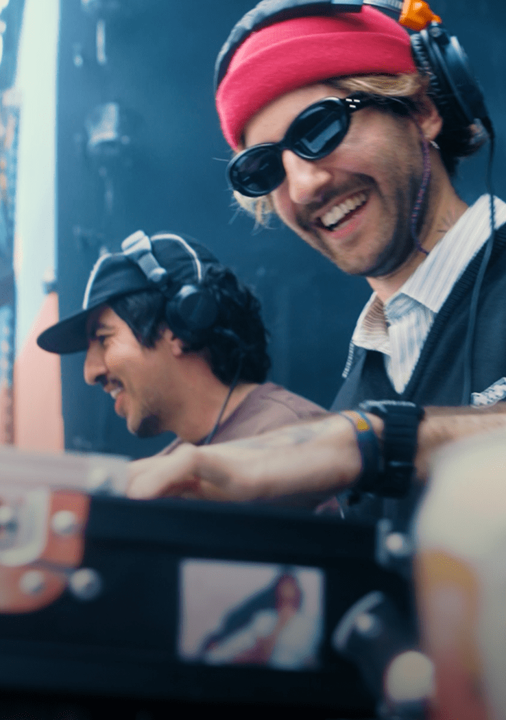 DJs sorrindo durante set ao vivo em evento, com headphones e mesa de som em primeiro plano
