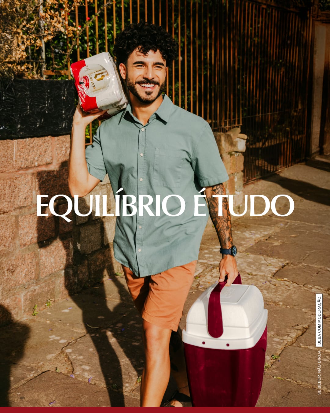 Homem sorridente carregando um pack de latas Bellavista no ombro e uma cooler vermelha e branca, caminhando por um pátio externo, com tagline "Equilíbrio é tudo"