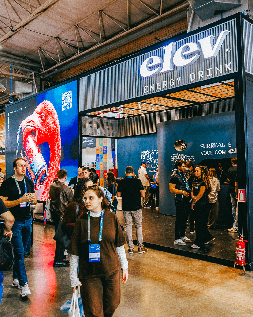 Estande da Elev Energy Drink em feira, com telão exibindo o anúncio do flamingo, letreiro "Surreal é você com elev" e público circulando pelo espaço