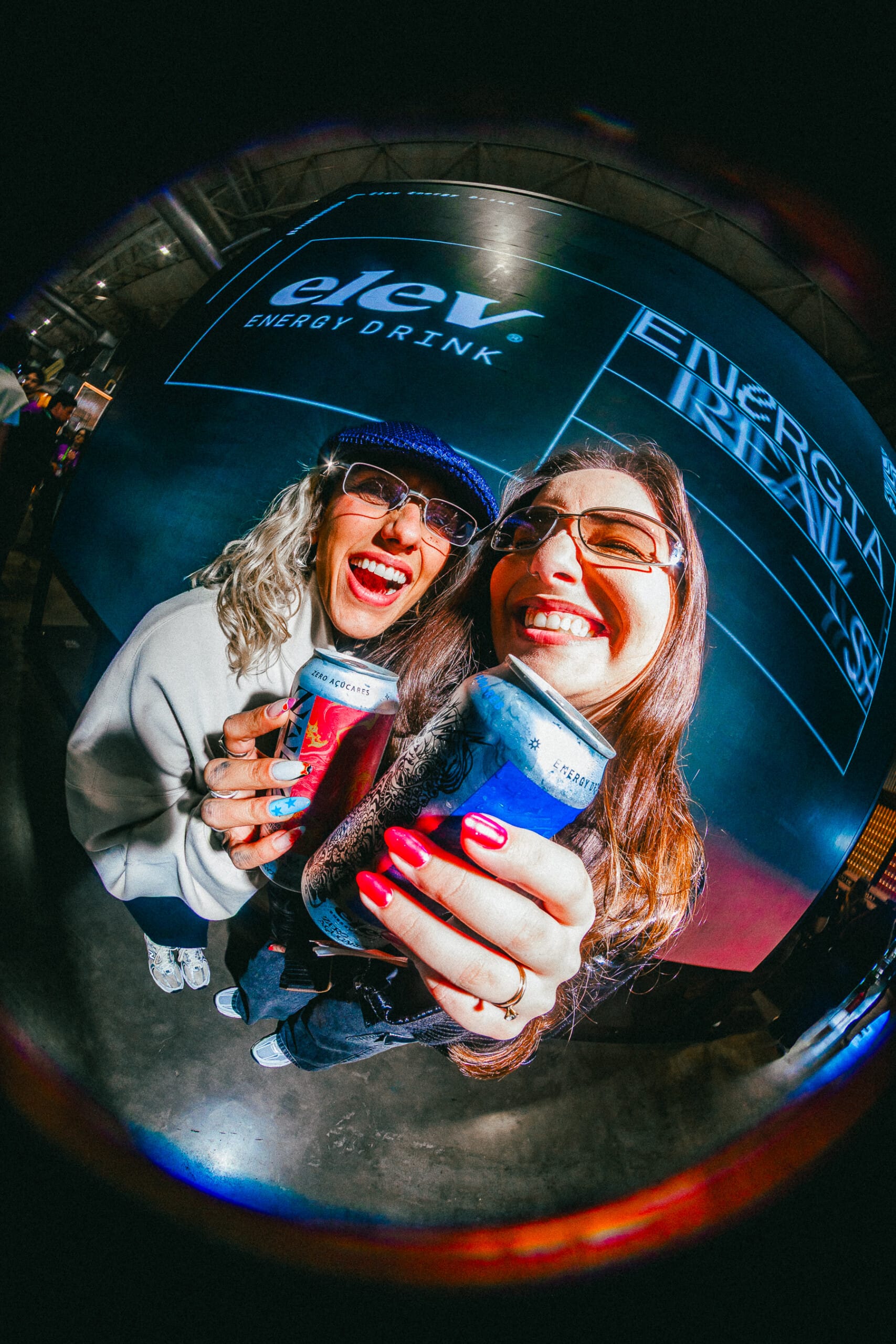 Selfie em lente olho-de-peixe de duas mulheres sorrindo, cada uma segurando uma lata Elev, com telão da marca ao fundo