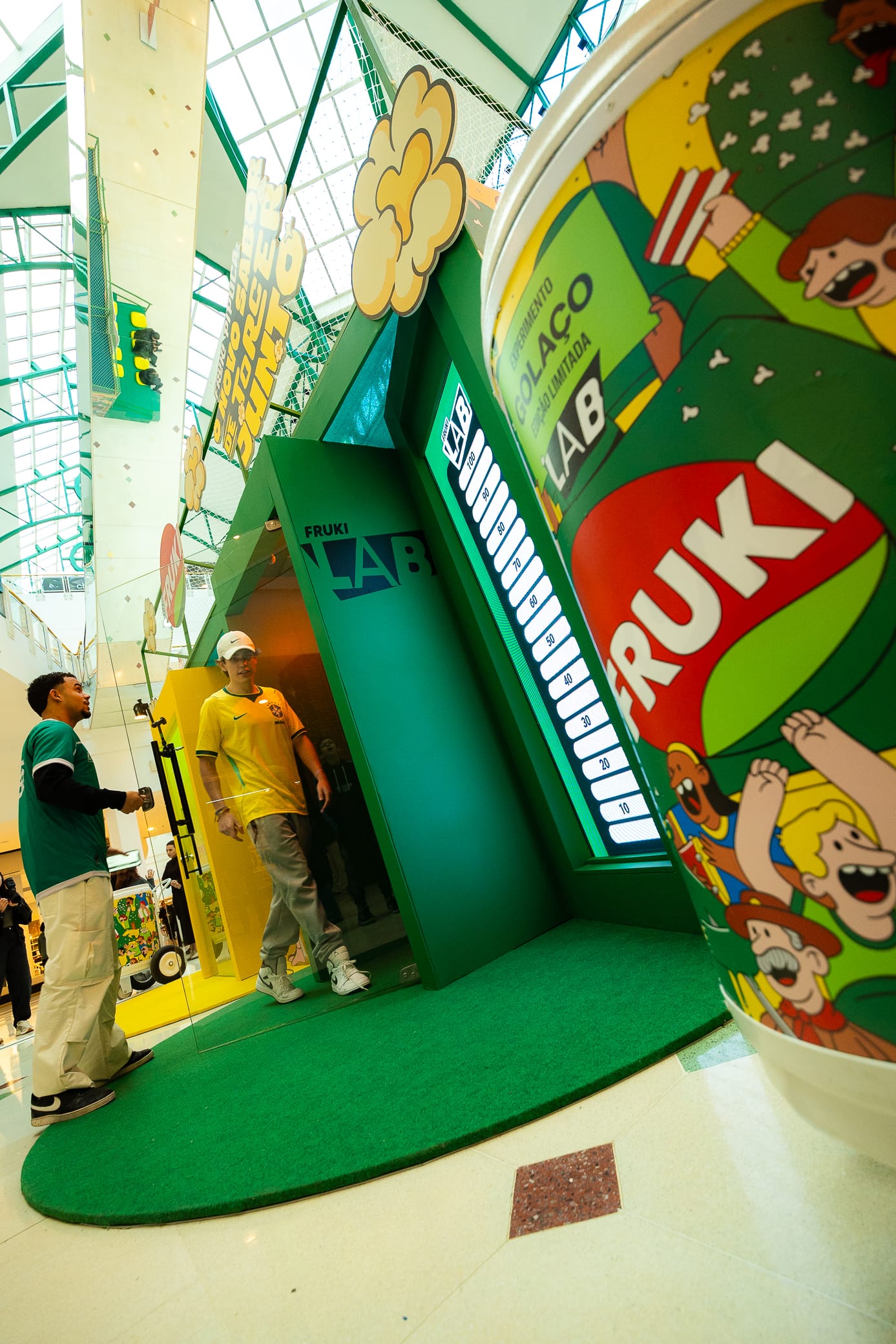 Cabine em formato de saco de pipoca da ativação "Fruki Lab" em shopping, com jovem de camisa do Brasil saindo do totem verde e amarelo, painel luminoso graduado de 10 a 100 e balde de pipoca gigante com a marca Fruki e arte do "Experimento Golaço Edição Limitada"