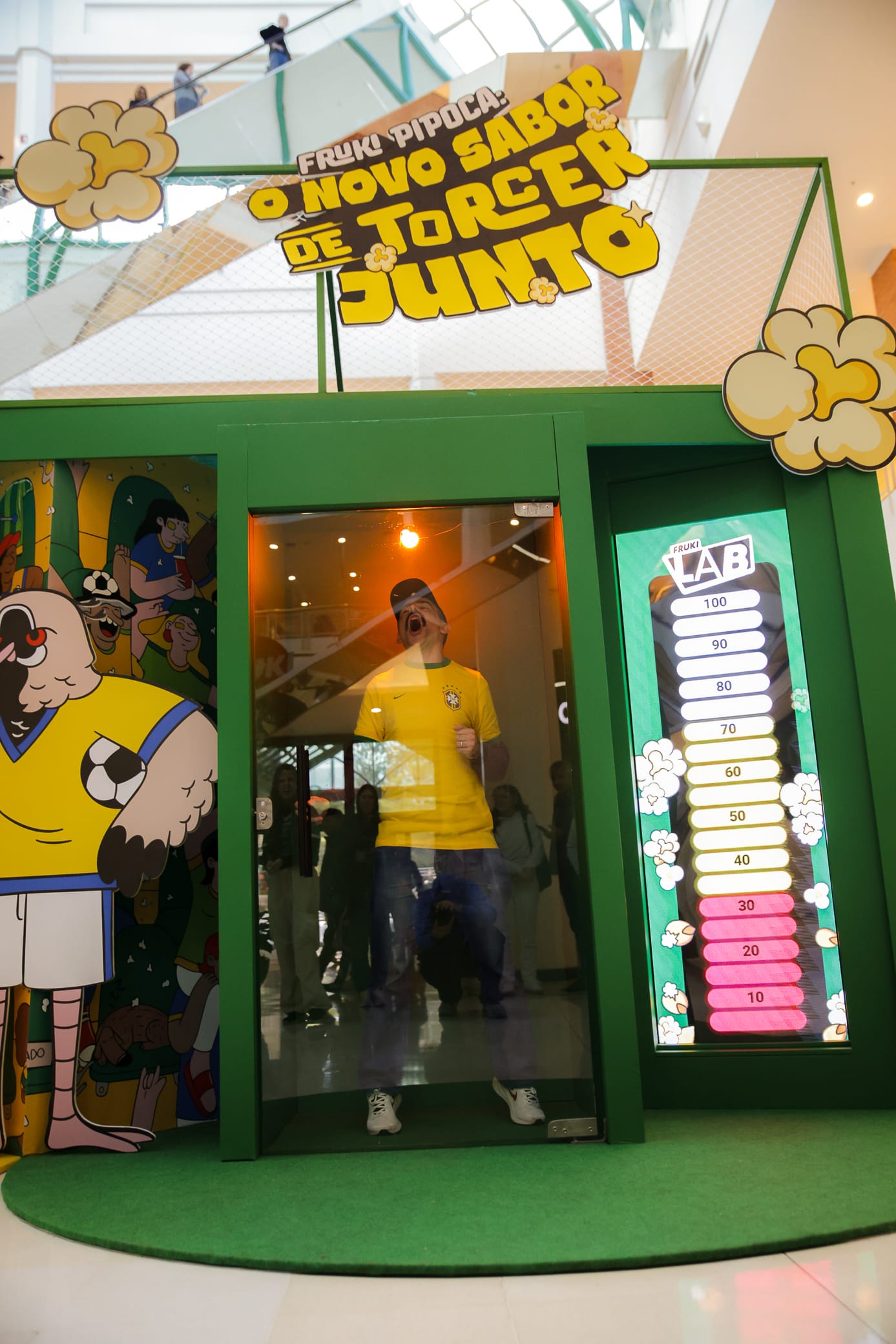Torcedor de camisa do Brasil comemorando dentro da cabine de vidro do "Fruki Lab", com letreiro "Fruki Pipoca: o novo sabor de torcer junto" e painel luminoso graduado de 10 a 100 ao lado